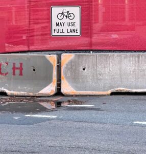 A sign that says bikes can use full lane, but the street is full of potholes and construction debris. This is a photo from Cambridge, Mass.